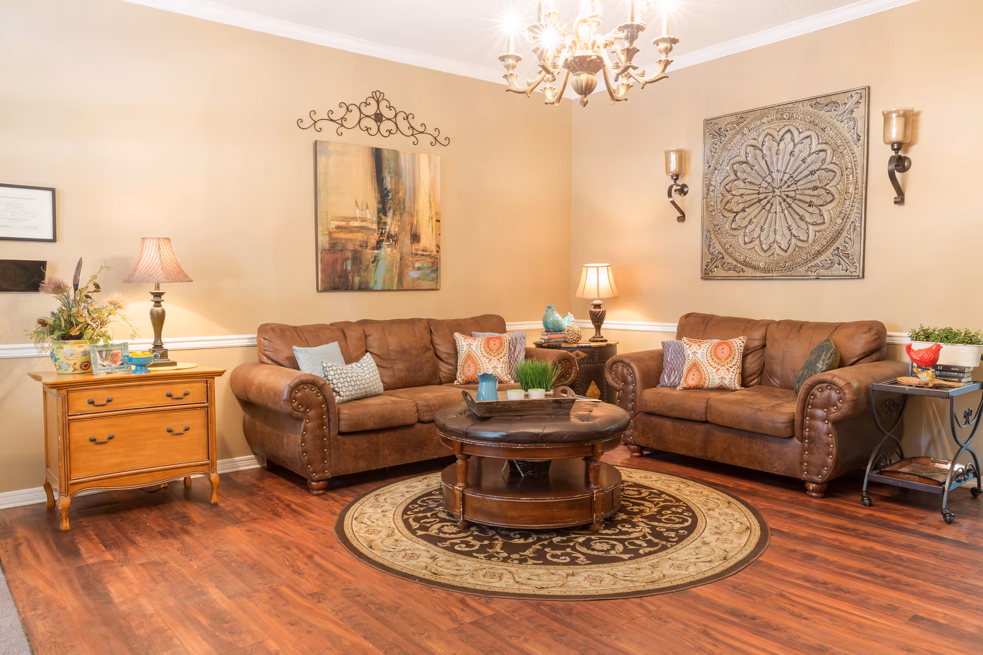A cozy living room with two brown leather sofas adorned with colorful patterned pillows. A round wooden coffee table with a tufted leather top sits on a circular patterned rug in the center. The walls are beige with decorative wall art and sconces. There are two side tables with lamps and decorative items, and a chandelier hanging from the ceiling. The floor is wooden.