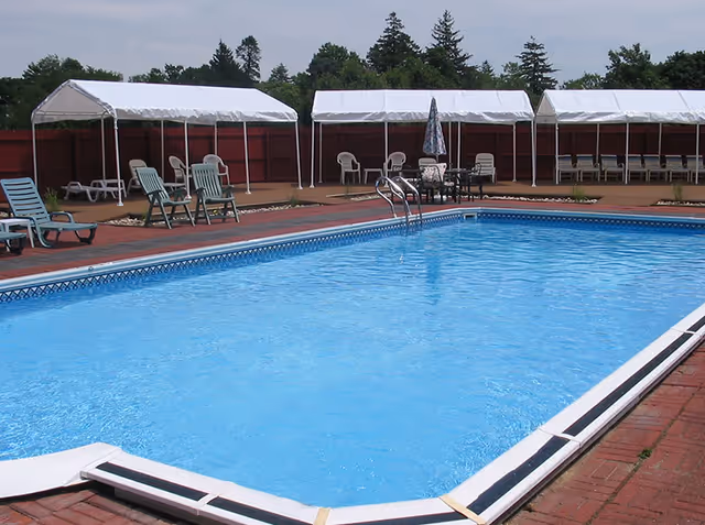 Outdoor swimming pool with clear blue water surrounded by a brick deck. Several lounge chairs and plastic chairs are arranged around the pool, with white canopy tents providing shade in the background. Trees and a red fence are visible behind the canopies.