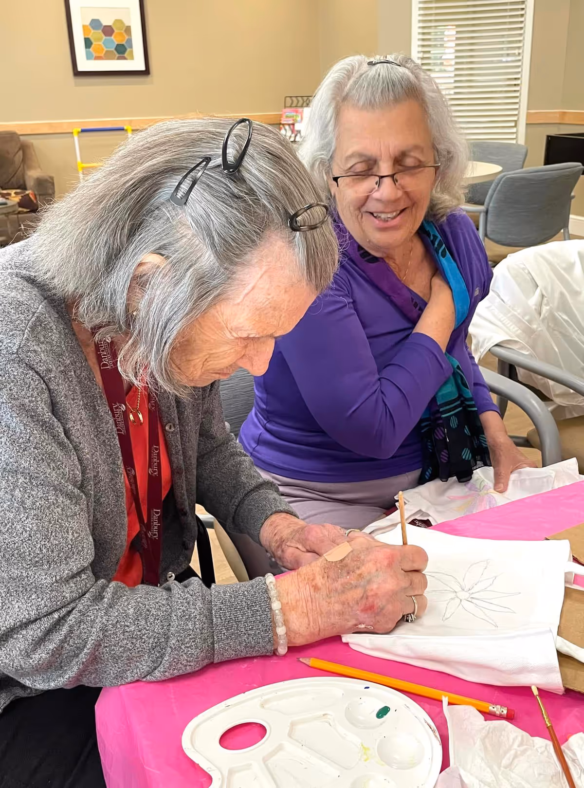 Two elderly women sitting at a table engaged in an art activity. One woman is drawing on a white cloth with a pencil, while the other woman watches and smiles. The table is covered with a pink tablecloth and has a paint palette and pencils on it. The room has chairs, a window with blinds, and a framed artwork on the wall.