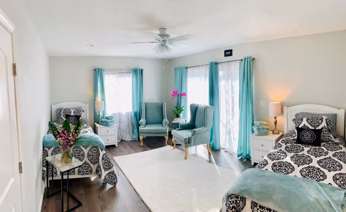 Bright twin bedroom with two beds, turquoise curtains, matching armchairs, nightstands, and floral accents.