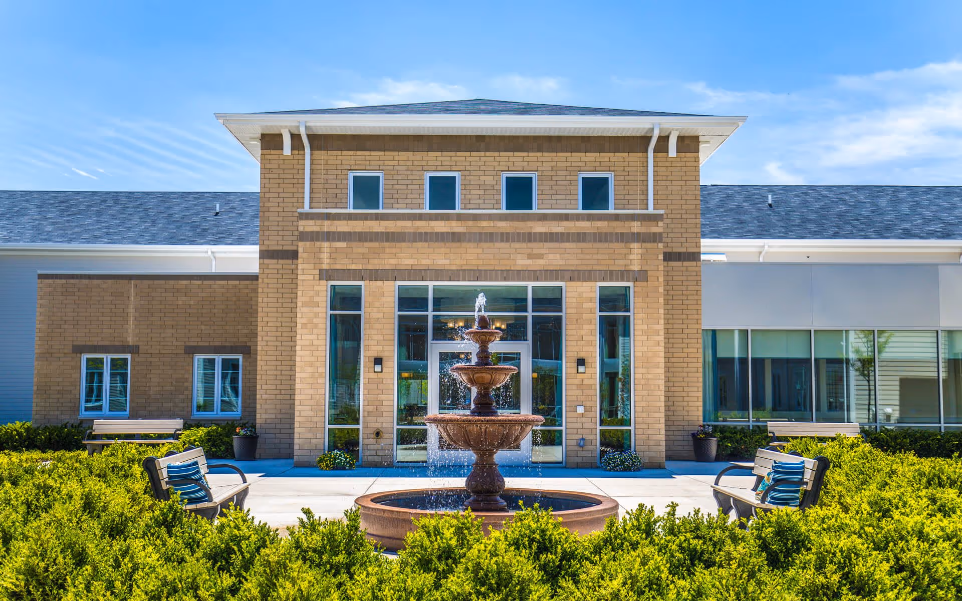 Front exterior view of a senior living facility named Ciel of Long Grove, featuring a three-tiered water fountain in the center, surrounded by green bushes and benches on either side under a clear blue sky.