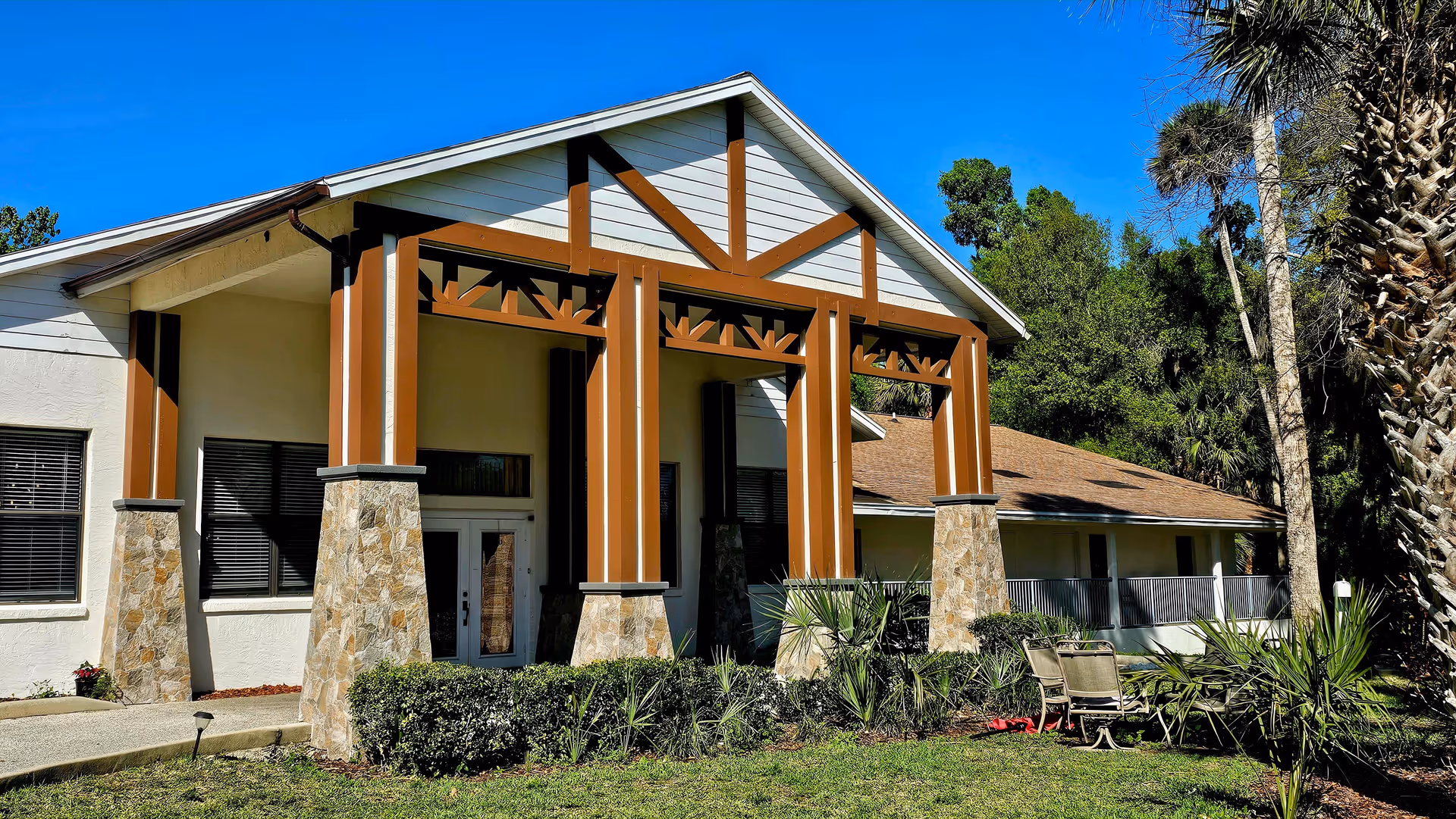 Exterior view of a building with a large covered entrance supported by stone and wooden pillars, surrounded by greenery and palm trees under a clear blue sky. There is outdoor seating with chairs and a table on the lawn.