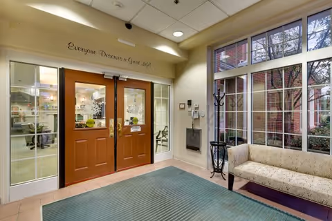 Entrance area of a senior living facility with double wooden doors featuring glass panels. Above the doors is a sign that reads 'Everyone Deserves a Great Life!'. To the right of the doors is a large window with a beige cushioned bench underneath. The floor has a greenish-blue rug and the walls are painted light yellow.