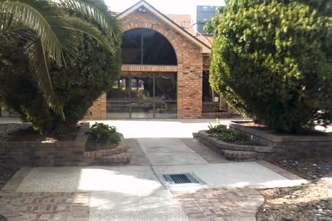 Front entrance of a brick building with an arched window, paved walkway and landscaped palms and trimmed shrubs.
