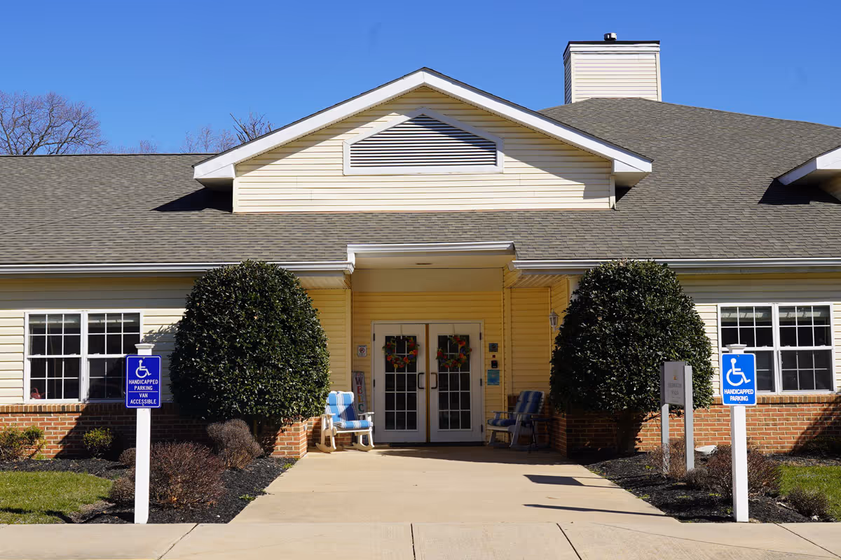 Front exterior view of a single-story building with a peaked roof and double glass doors decorated with wreaths. Two large trimmed bushes flank the entrance, and there are two blue handicapped parking signs on white posts in front of the building. Two chairs with cushions are placed near the entrance on either side. The sky is clear and blue.