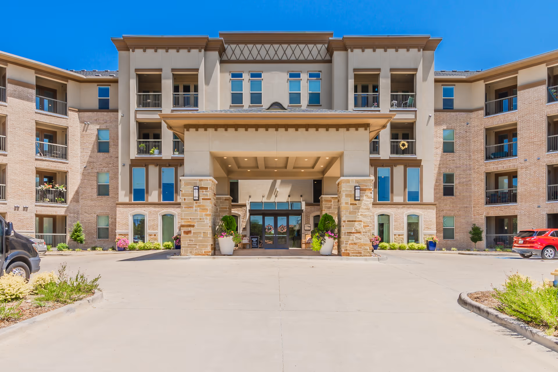 Front exterior view of a multi-story senior living facility building with a covered entrance supported by stone pillars, potted plants, balconies, and clear blue sky.