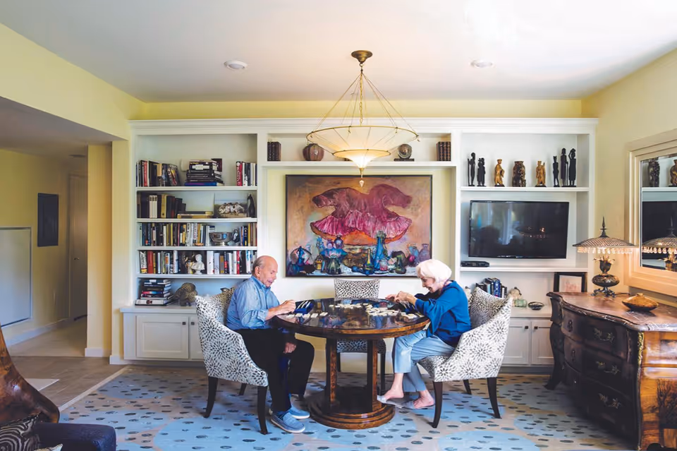 Two older adults sit at a round table in a bright sitting room with built-in bookshelves, artwork, and a TV.