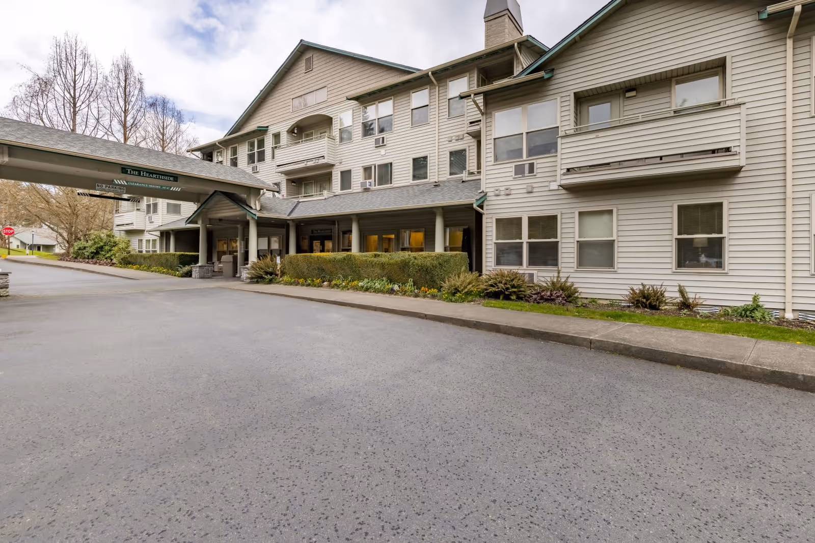 Exterior view of a multi-story senior living facility building with beige siding, multiple windows, balconies, and a covered entrance labeled 'The Hearthside'. There are shrubs and small plants along the building and a paved driveway in front.