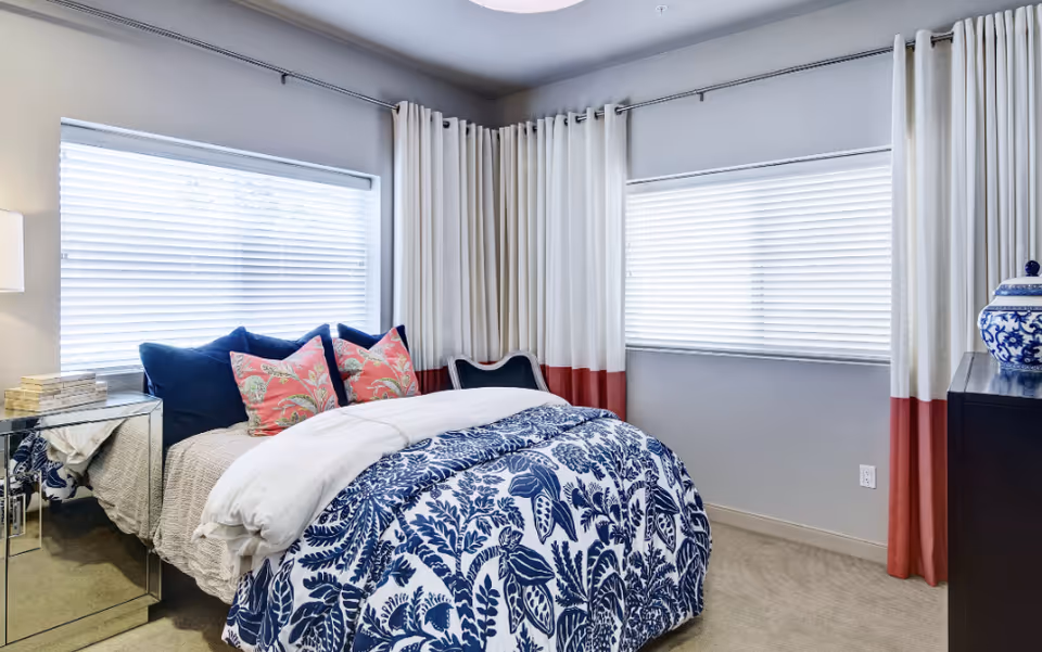 Bright bedroom with blue-white botanical comforter, pink pillows, and large windows.