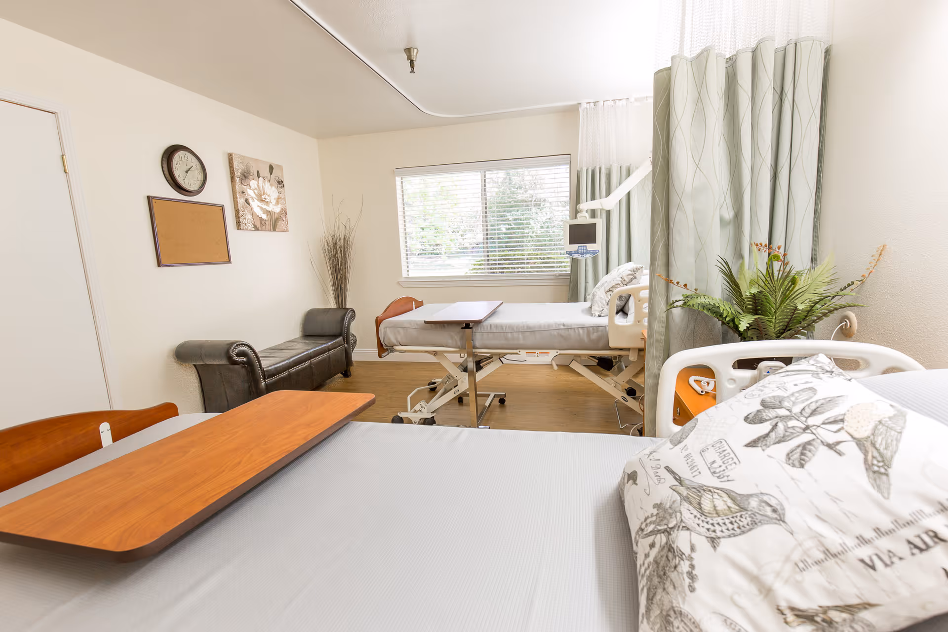 A bright and clean patient room with two hospital beds separated by a privacy curtain. One bed is in the foreground with a pillow featuring bird illustrations, and the other bed is near a window with blinds. There is a wooden overbed table on the bed in the foreground, a small black bench against the wall, a clock, a corkboard, and a floral wall decoration. A potted plant is placed near the curtain.