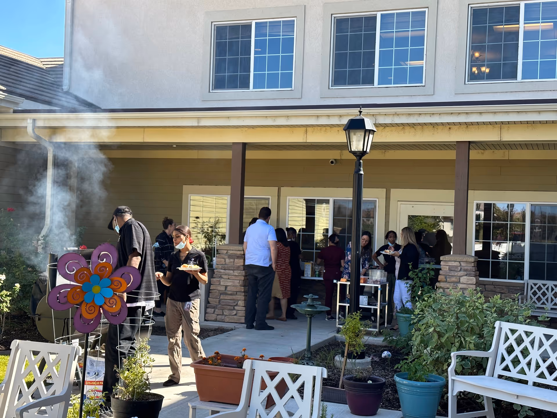 Outdoor patio area at Wingfield Skilled Nursing And Rehabilitation Center with several people gathered near a covered porch. Some individuals are standing by a grill with smoke rising, while others are socializing near tables. The area has garden plants, white benches, and a decorative flower stake.