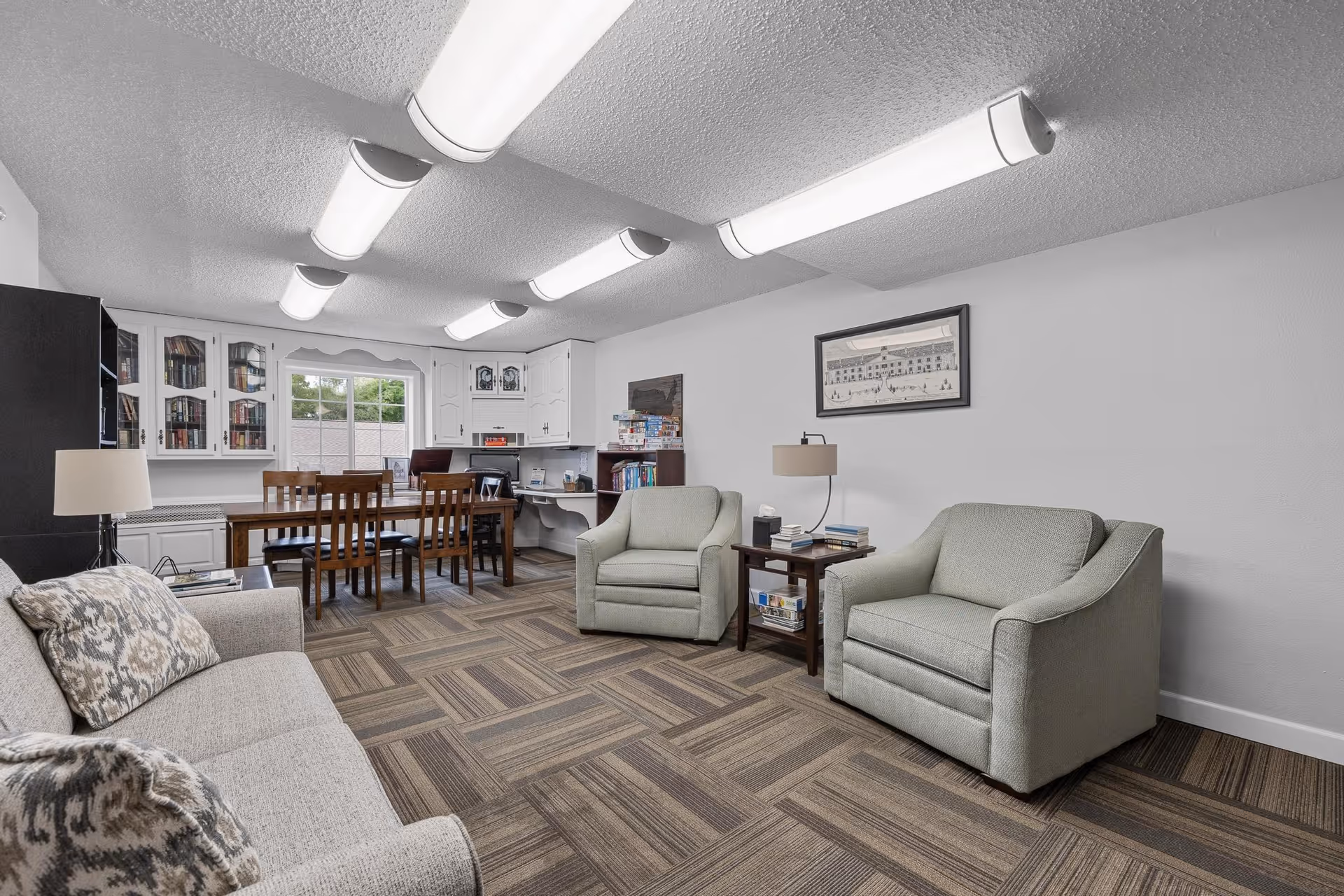 A well-lit common area with a beige sofa and two light green armchairs arranged around a wooden side table with a lamp and books. In the background, there is a wooden dining table with chairs, a window, and white cabinets with glass doors containing books. The room has a patterned carpet and fluorescent ceiling lights.