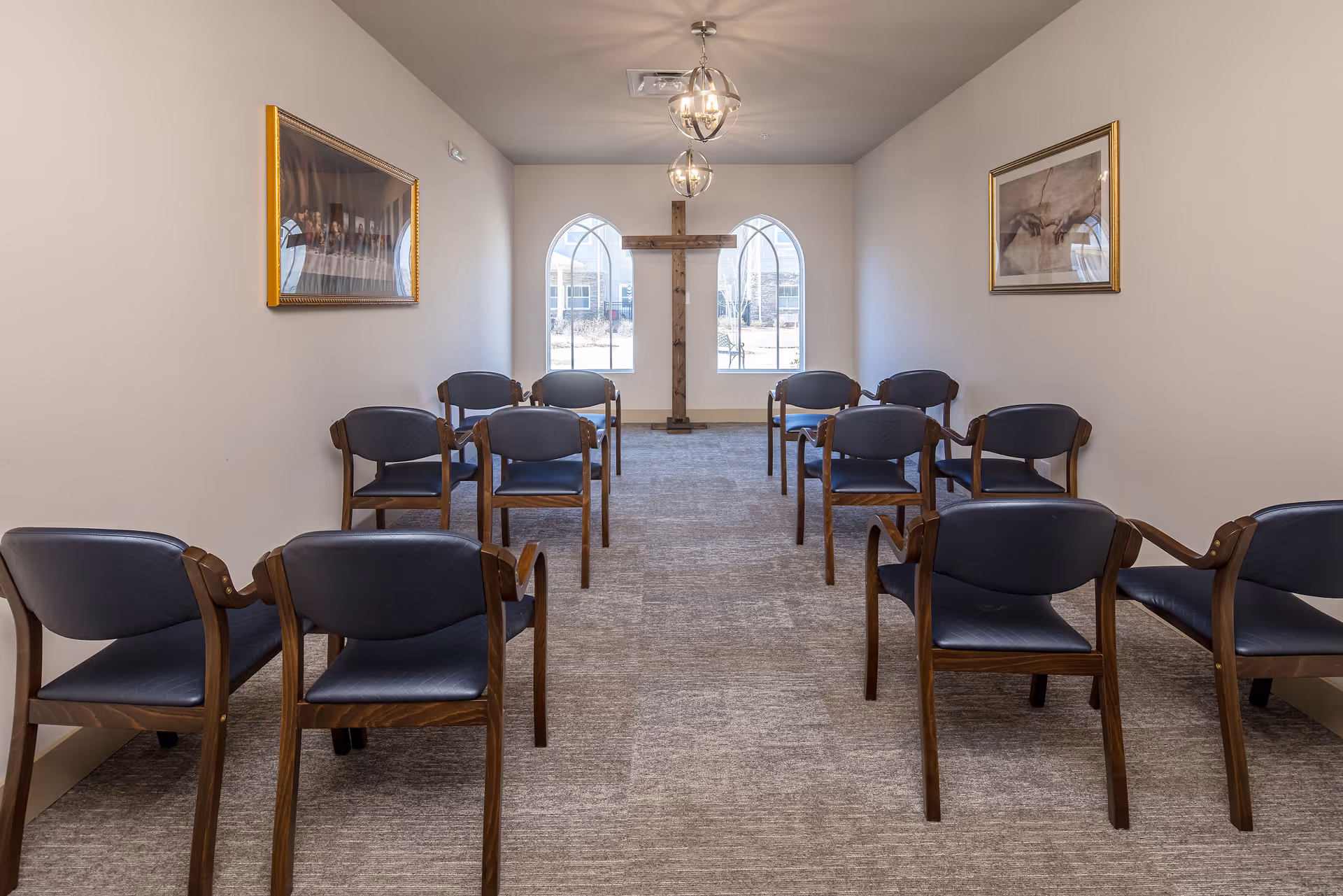 Small chapel-style meeting room with rows of chairs facing a wooden cross and arched windows.