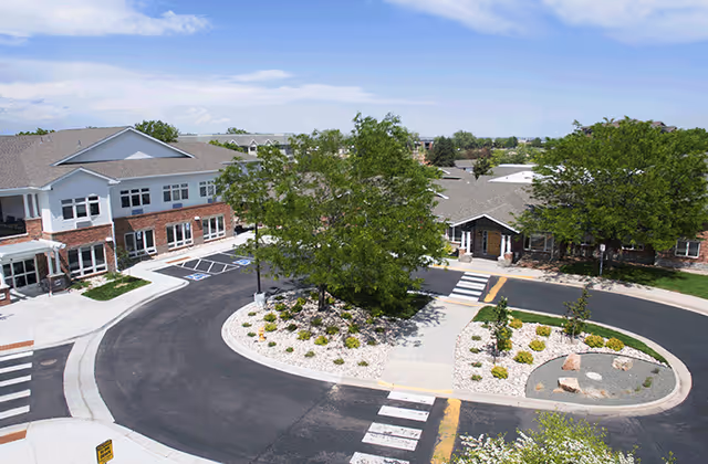 Aerial view of the entrance to New Mercer Commons Assisted Living facility showing a circular driveway with landscaped islands featuring trees and shrubs, surrounded by two-story brick and siding buildings under a partly cloudy sky.