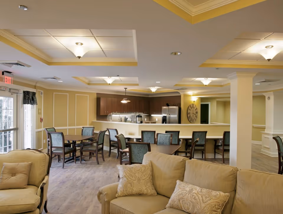 A spacious and well-lit common area in a senior living facility featuring a beige sofa with patterned cushions in the foreground, several wooden tables and chairs arranged for dining or socializing, and a kitchen area with dark wood cabinets, a stainless steel refrigerator, and a large wall clock. The room has light yellow walls, recessed ceiling lights, and a door with an exit sign.