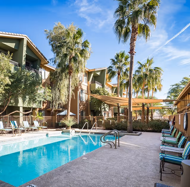 Outdoor swimming pool and lounge area with palm trees next to a three-story residential building under a blue sky.