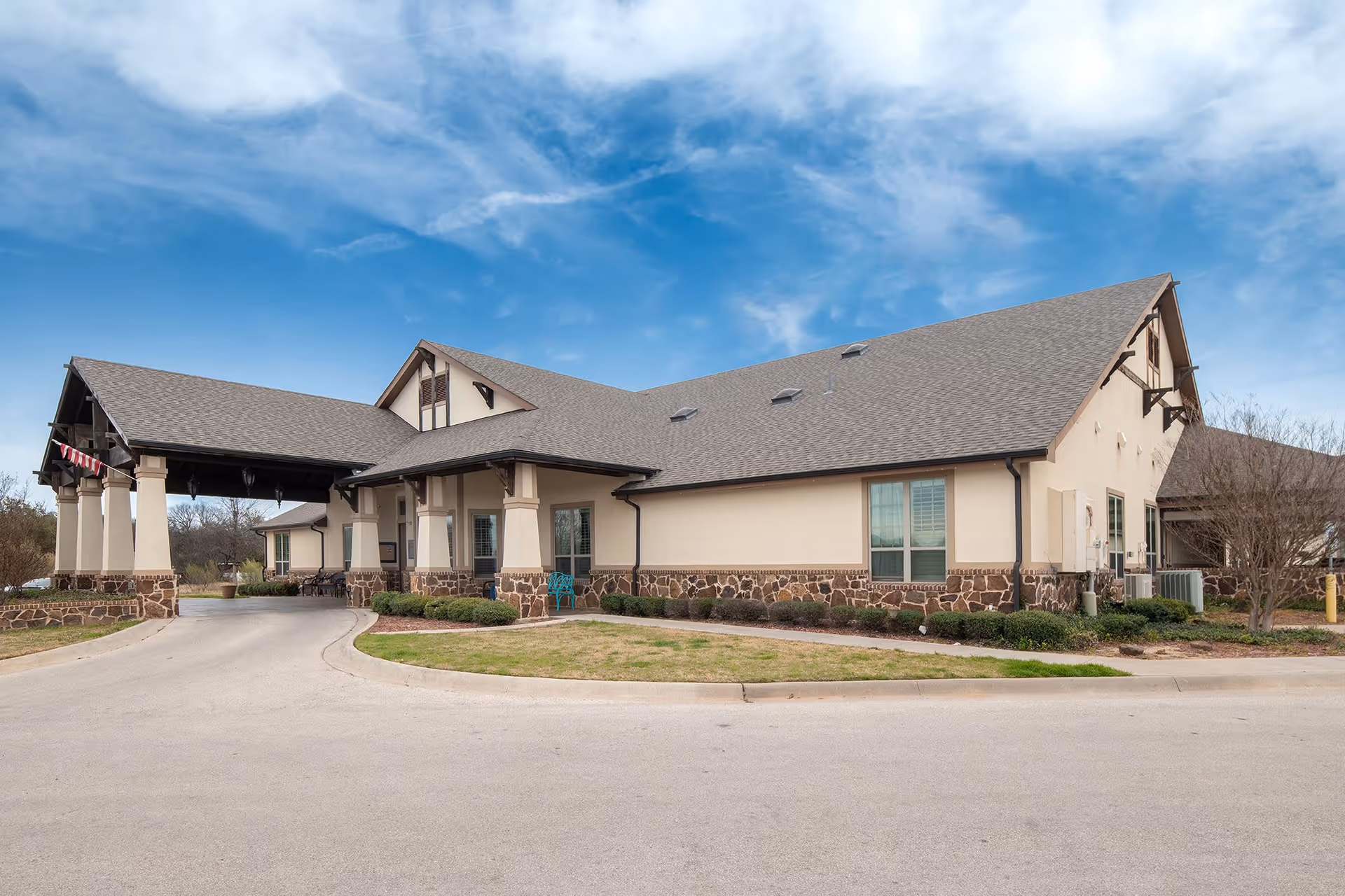 Exterior view of a single-story senior living facility building with a covered entrance, beige walls with stone accents at the base, multiple windows, and a gray shingled roof under a partly cloudy blue sky.
