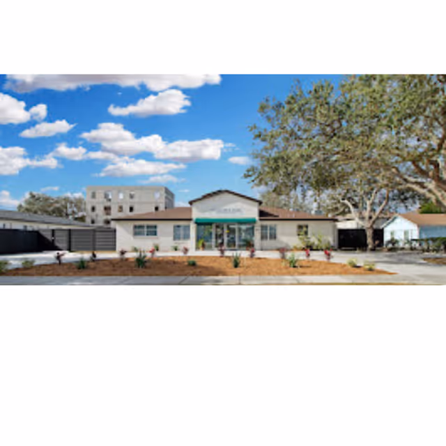 Single-story assisted living building with a central glass entrance under a green awning, landscaped front beds, and a large tree against a blue sky.