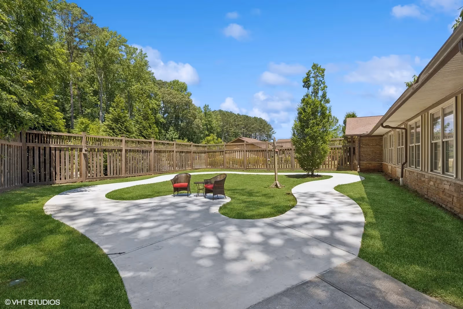 Outdoor courtyard area with a curved concrete pathway surrounding a grassy lawn. Two wicker chairs with red cushions and a small table are placed on the grass. The area is enclosed by a wooden fence and bordered by trees and a building with multiple windows. The sky is blue with some clouds.