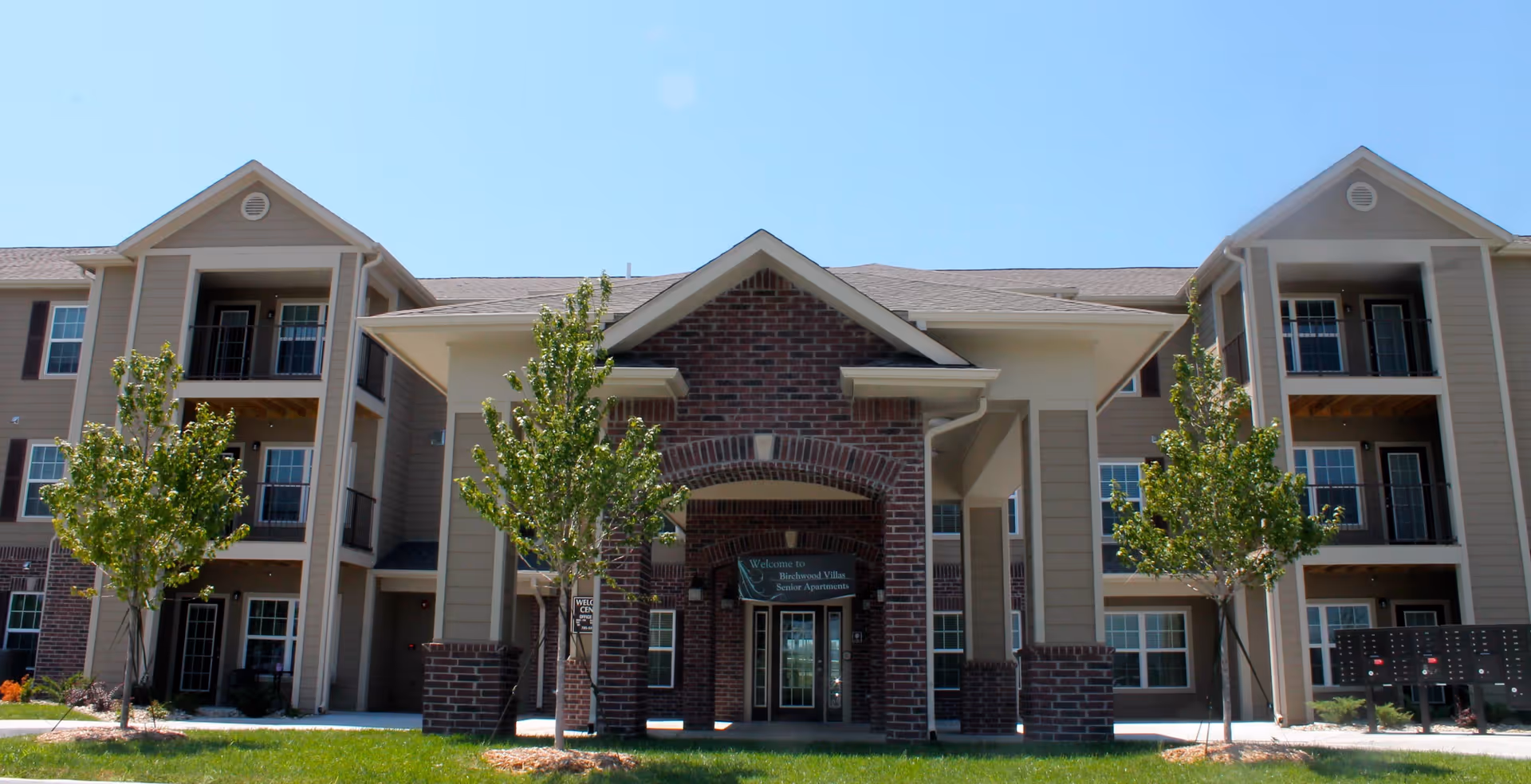 Front exterior view of Birchwood Villas Senior Apartments building with a brick and beige facade, multiple windows, balconies, and a covered entrance. There are small trees and green grass in front of the building under a clear blue sky.