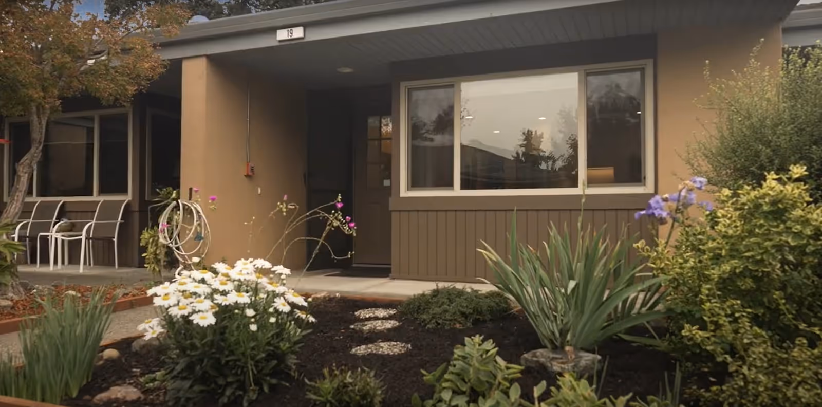 Exterior view of a single-story building entrance with a large window and a door under a covered porch. There are various plants and flowers, including white daisies and purple flowers, in a landscaped garden bed in front of the building. Several chairs are visible on the left side near another window.