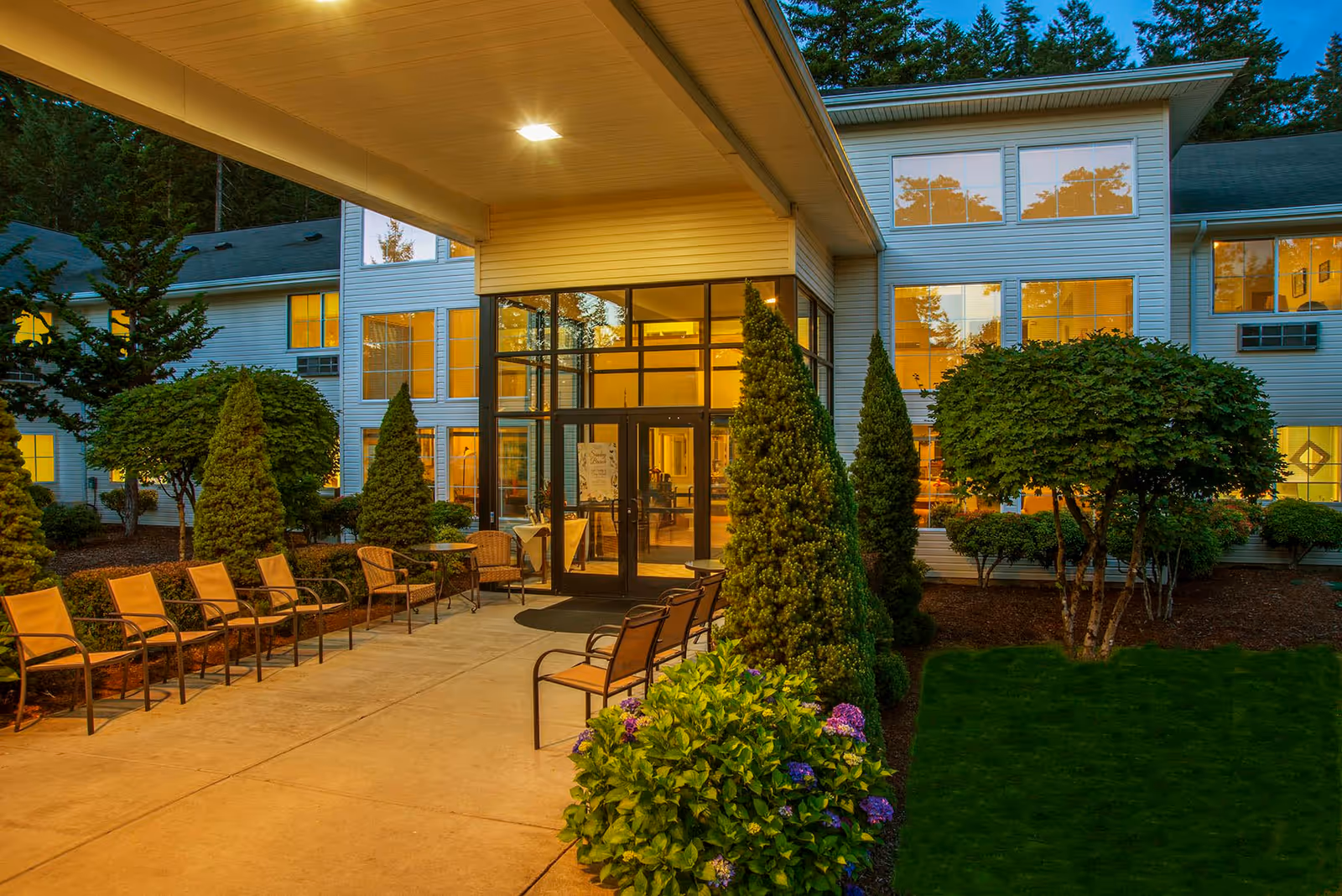Entrance of a senior living facility at dusk with a covered walkway, outdoor seating with chairs and tables, well-maintained landscaping including bushes, trees, and flowers, and large windows showing warm interior lighting.