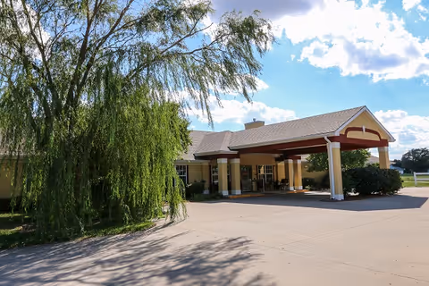 Front entrance of a single-story senior living building with a covered porte-cochere, driveway, and a large weeping willow tree nearby.