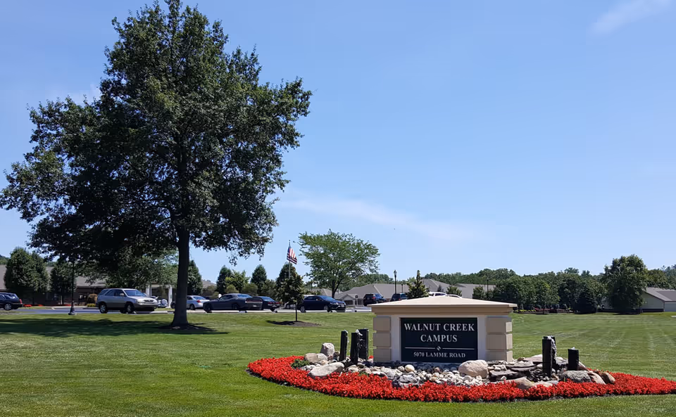 A landscaped outdoor area with a large tree, green grass, and a flower bed with red flowers surrounding a stone sign that reads 'Walnut Creek Campus 5070 Lamme Road'. Several cars are parked in the background under a clear blue sky.