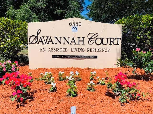 Entrance sign reading "Savannah Court, An Assisted Living Residence" with address 6550, set among mulch and flowering plants.