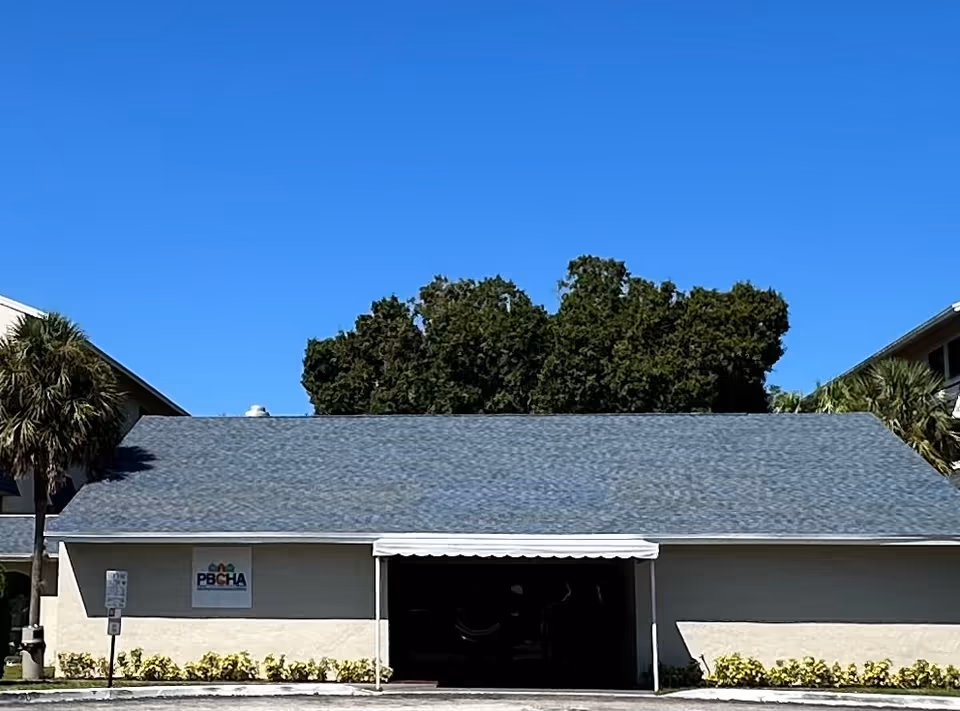 Exterior view of a single-story building with a gray roof and small awning, landscaping along the front, and a clear blue sky.