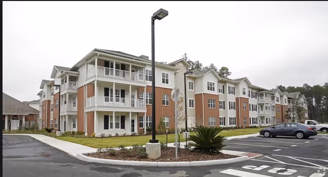 Three-story senior living building with covered balconies, landscaped grounds and a parking lot in front.