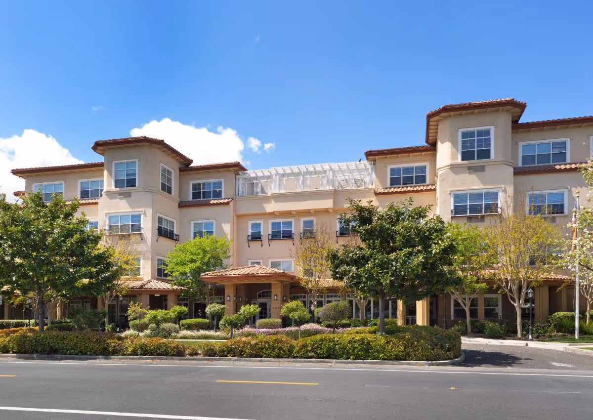 Exterior view of a multi-story senior living facility building with beige walls and red-tiled roofs, surrounded by green trees and landscaped bushes under a clear blue sky.
