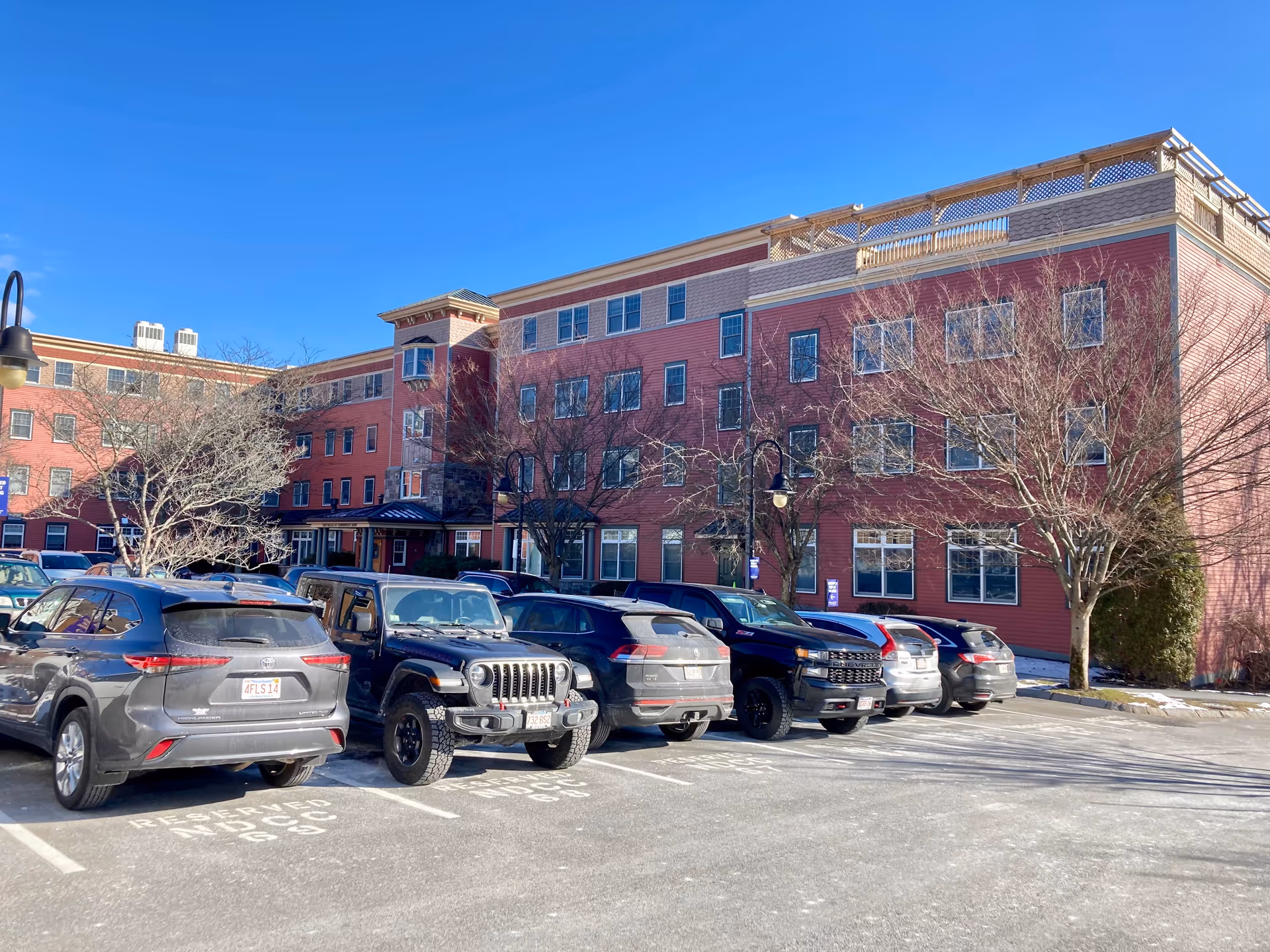 Exterior view of a multi-story senior living facility building with red siding and multiple windows. Several cars are parked in the parking lot in front of the building under a clear blue sky. Leafless trees are visible near the building.