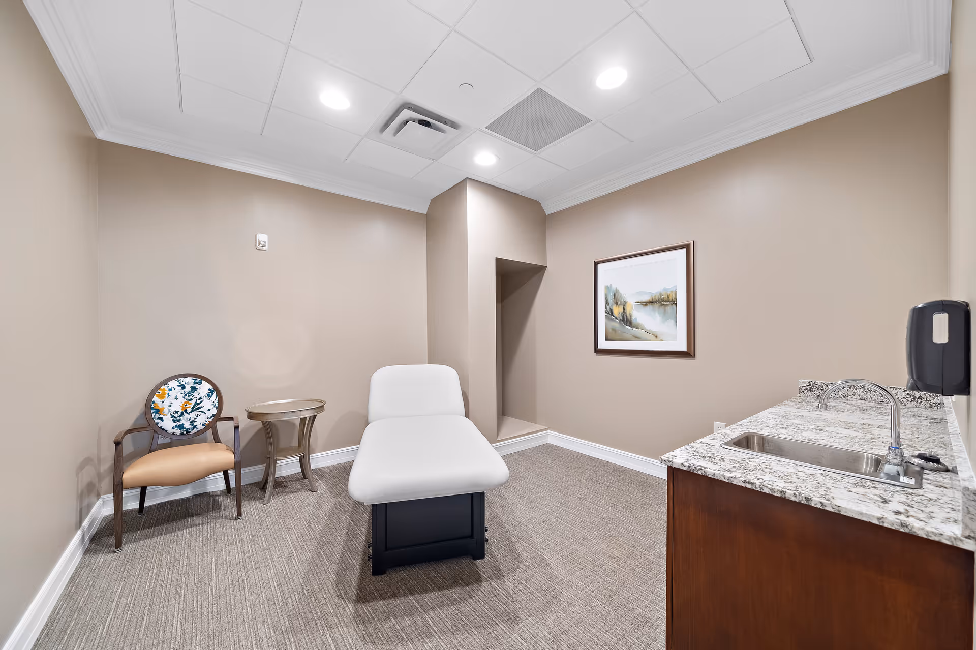Neutral-toned treatment room with an exam table, a chair and side table, framed artwork, and a sink countertop.
