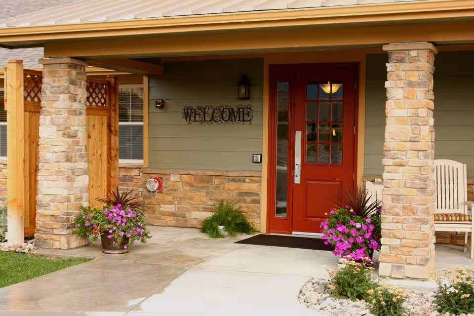 Front entrance porch with a red door, stone columns, potted flowers and a 'WELCOME' sign.
