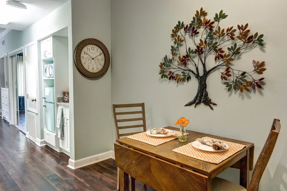 A small dining area with a wooden table set for two, featuring orange patterned placemats, plates, and napkins. A small vase with orange flowers is centered on the table. Two wooden chairs are positioned at the table. On the wall above the table is a decorative metal tree sculpture with multicolored leaves. To the left, a hallway leads to other rooms, and a round wall clock with a woven frame is mounted on the wall. The floor is dark wood, and the walls are painted light gray.