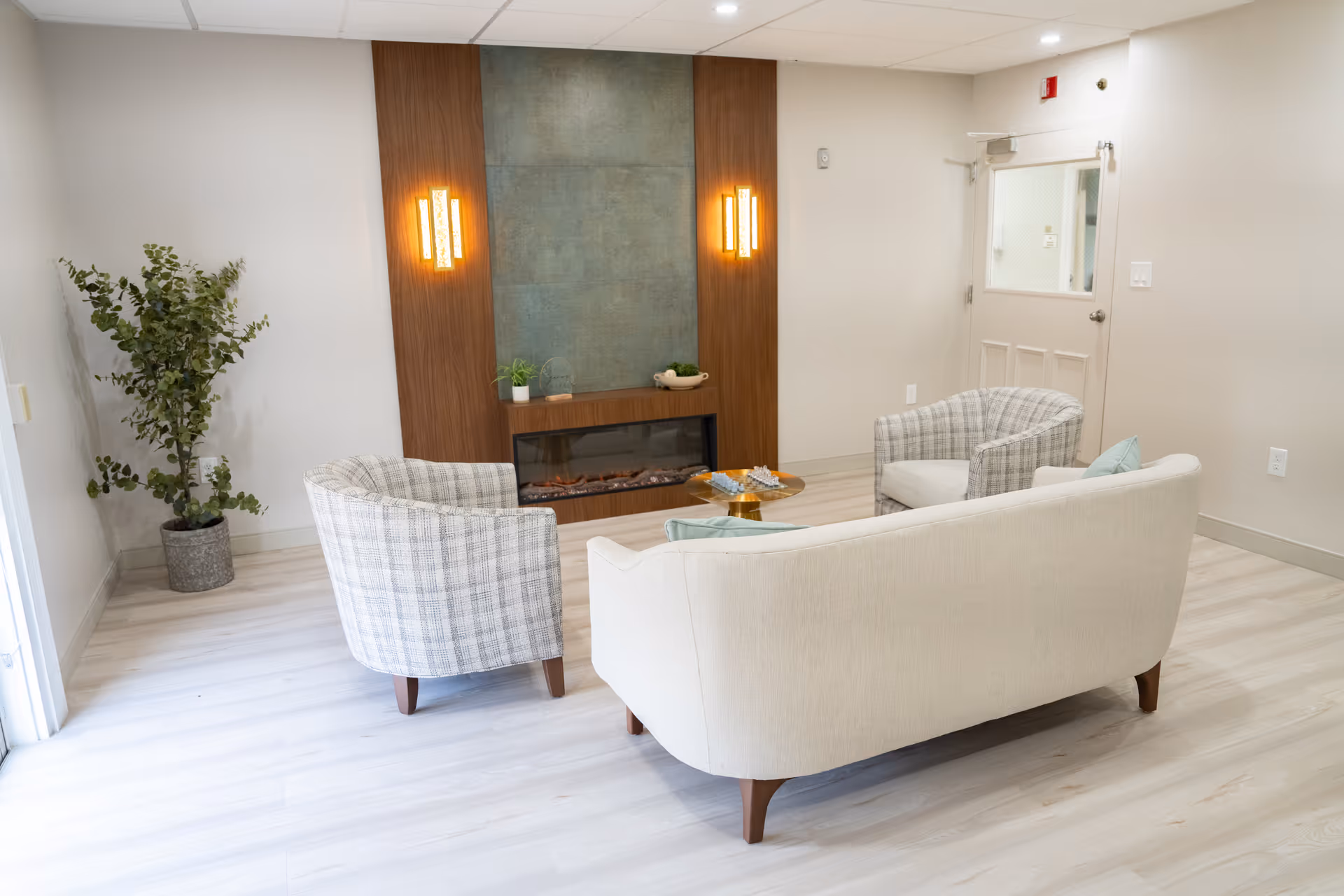 A cozy sitting area with a beige sofa and two plaid armchairs arranged around a small round wooden table. The room features light wood flooring, a modern electric fireplace set into a wooden and stone accent wall, two vertical wall sconces emitting warm light, a potted plant in the corner, and a white door with a window on the right side.