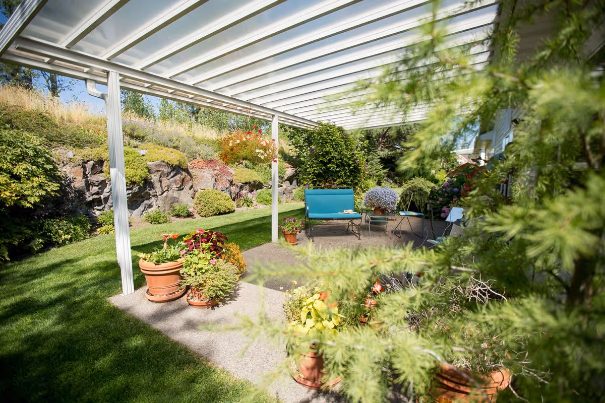 Covered patio with potted plants, a turquoise bench and small table under a white pergola overlooking a lawn and rock garden.