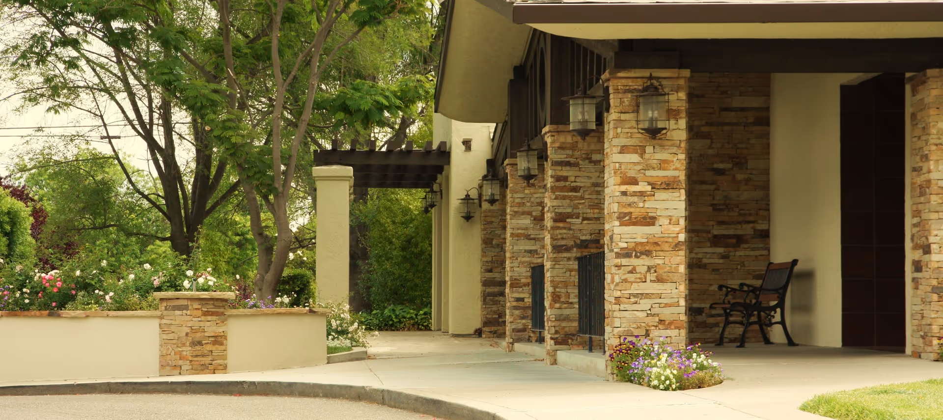 Covered entrance of a stone-clad building with columns, a bench, and landscaped flowers and trees.
