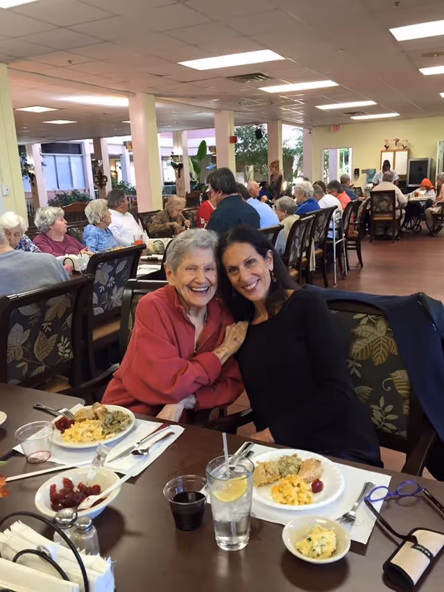 Two women smiling and sitting closely together at a dining table in a senior living facility. The table is set with plates of food including macaroni and cheese, stuffing, and cranberry sauce. In the background, many elderly people are seated at tables in a large, well-lit dining room with light-colored walls and ceiling lights.