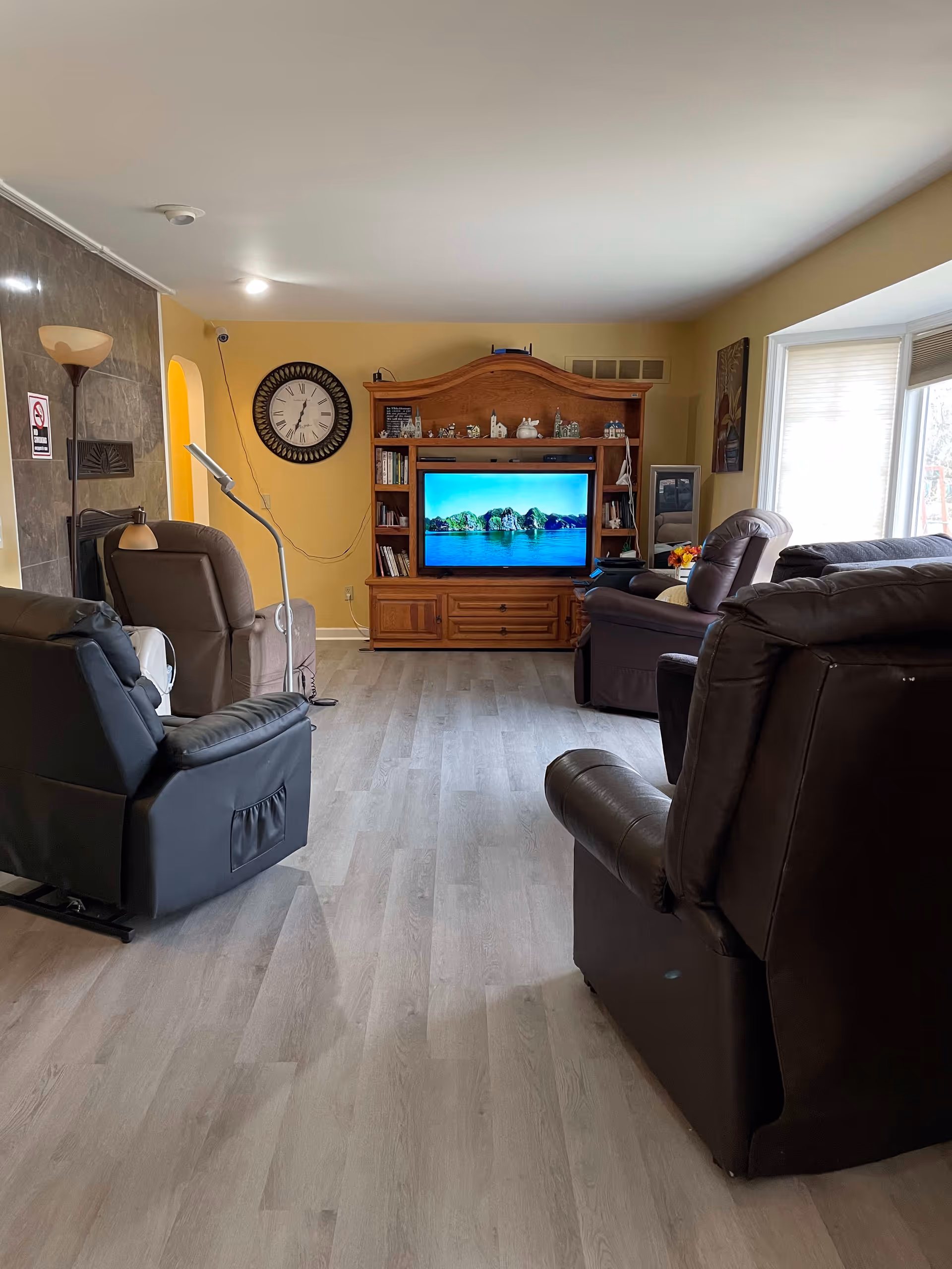 A cozy living room with several recliner chairs arranged facing a wooden entertainment center with a flat-screen TV displaying a scenic image of water and trees. The room has light-colored wood flooring, a large wall clock, a floor lamp, and a large window letting in natural light.