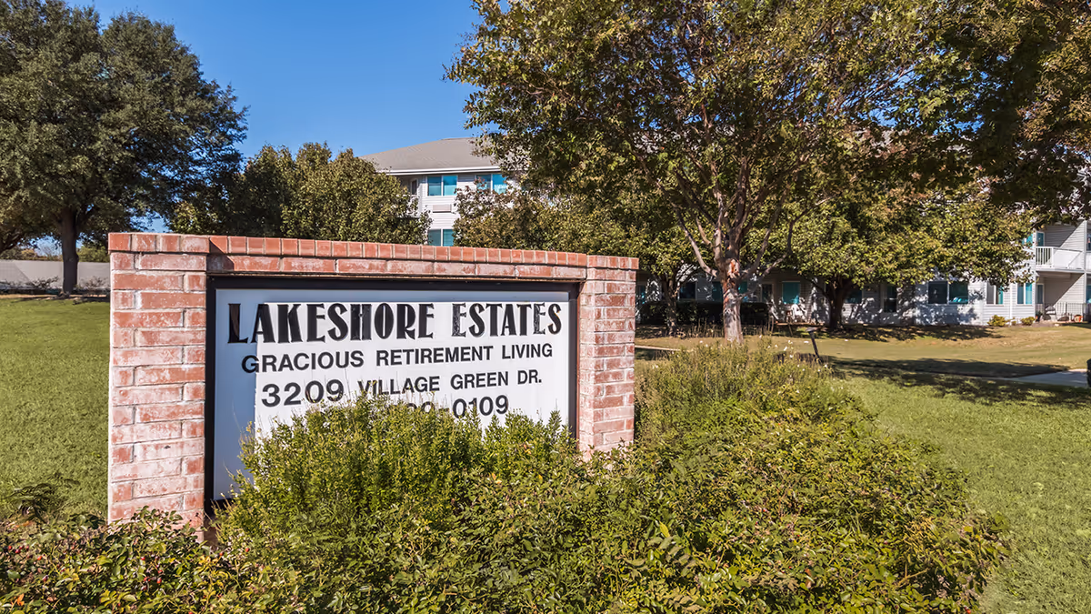 Brick entrance sign reading "Lakeshore Estates Gracious Retirement Living" on a grassy lawn with trees and the facility building visible behind it.