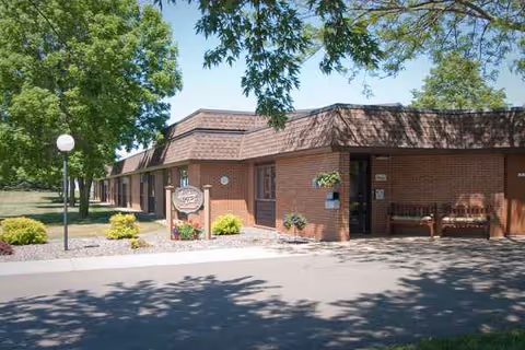 Brick senior living facility exterior with entrance, landscaping, and a shaded sidewalk.