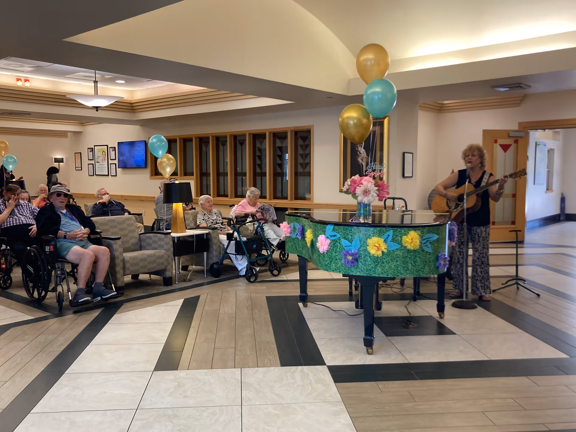 A group of elderly people seated in a common area of a senior living facility, some in wheelchairs, watching a woman playing guitar and singing near a decorated piano with balloons and flowers.