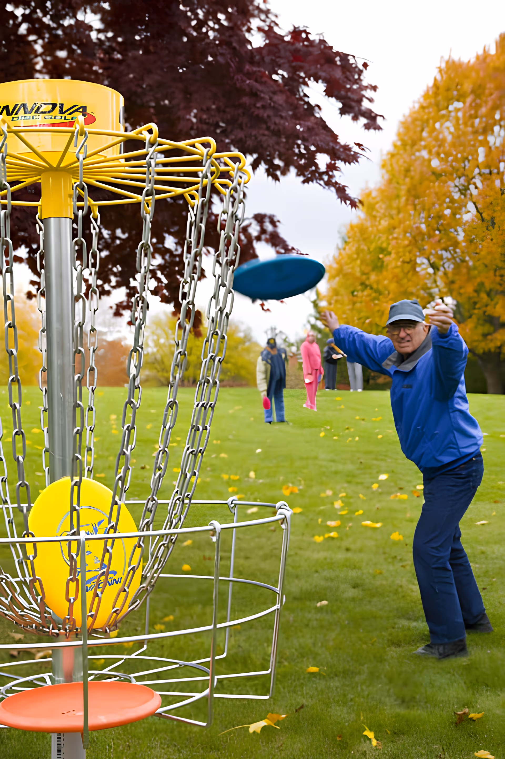 An elderly man wearing a blue jacket and cap is playing disc golf outdoors on a grassy area with autumn-colored trees in the background. A yellow disc golf basket with chains and discs is in the foreground.