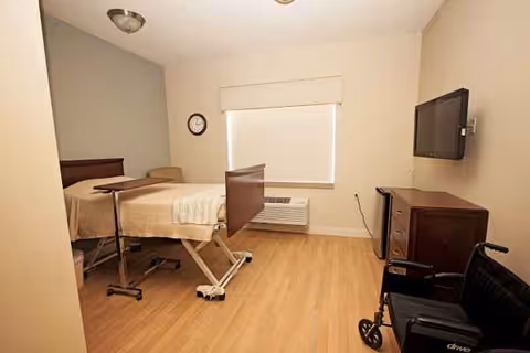A hospital-style patient bedroom with adjustable bed, dresser, wheelchair, and wall-mounted TV.
