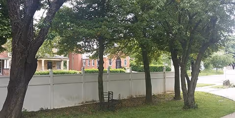 A grassy yard with several trees, a white privacy fence, and a small metal bench beside a sidewalk with houses across the street.