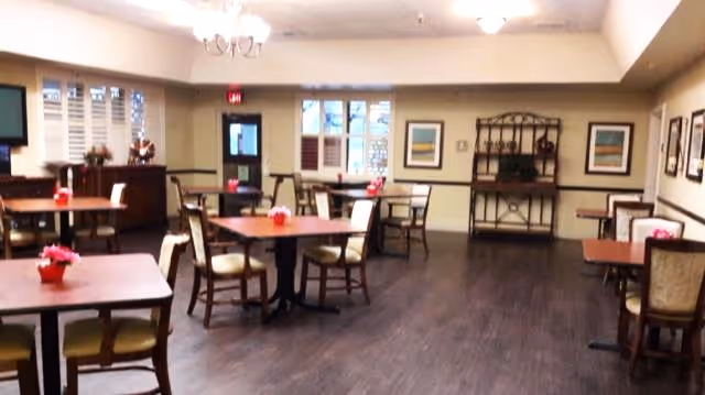 A communal dining room with multiple small tables and chairs, dark wood flooring, and a serving sideboard.