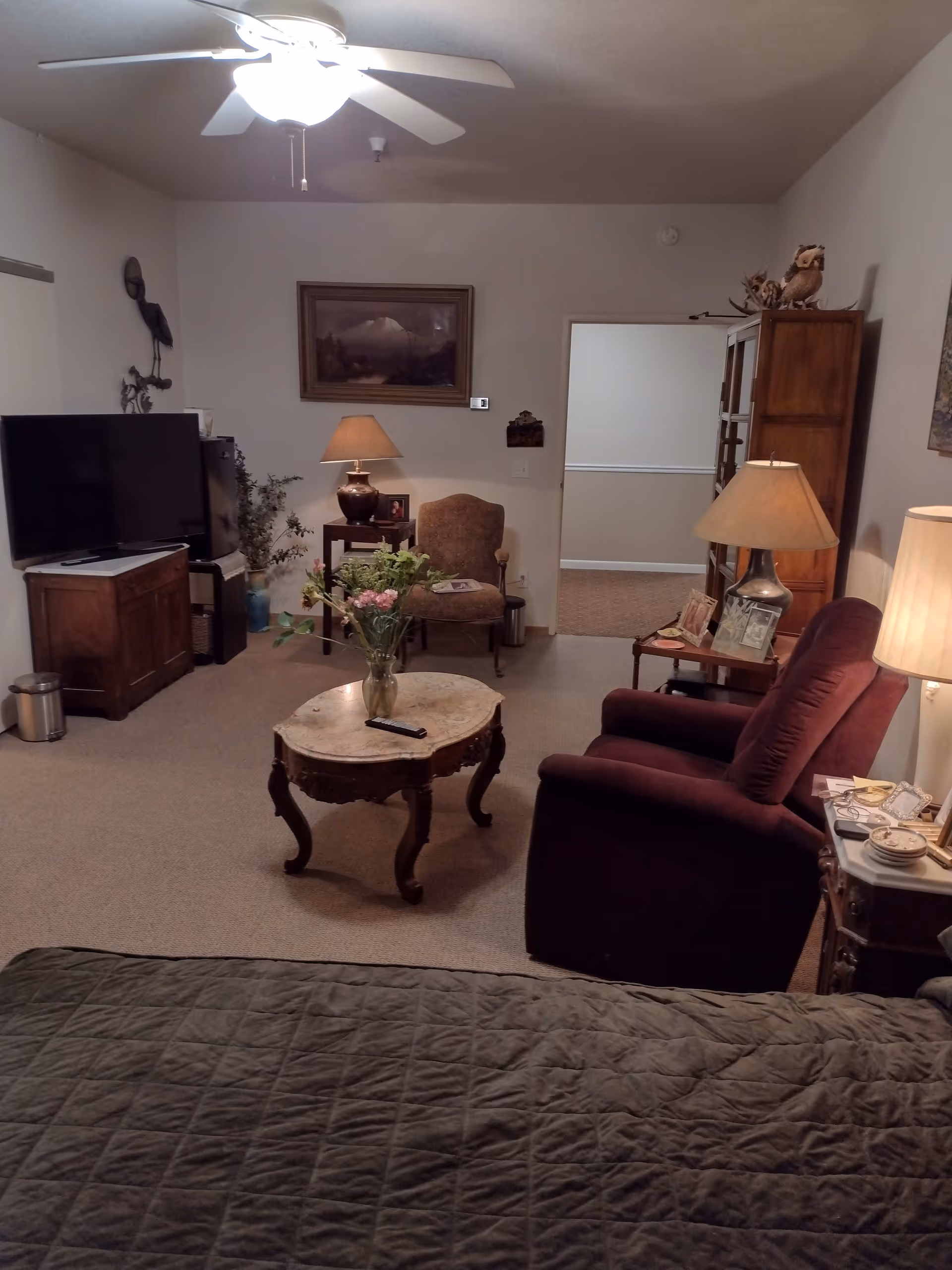A cozy living room area in an assisted living facility featuring a ceiling fan with light, a flat-screen TV on a wooden stand, a small wooden table with a lamp and a chair, a coffee table with a vase of flowers, a plush armchair, and a side table with a lamp and framed photos. The room has carpeted flooring and neutral-colored walls with framed artwork.