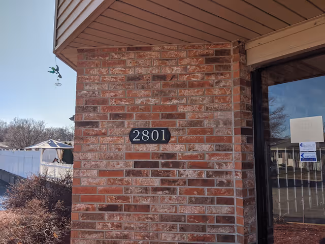 Close-up view of a brick wall corner of a building with the number 2801 displayed on a black plaque. To the right is a glass door with some notices posted on it. In the background, there is a white fence, some bushes, and a clear blue sky.
